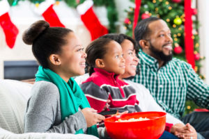 Happy couple and their teenage daughter and elementary age son watch a Christmas movie together during the Christmas season. The girl is holding a bowl of popcorn and the remote control. They are all smiling while watching the movie. A Christmas tree, stockings and a fireplace are in the background.